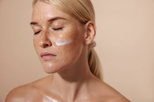 Close-up of a woman with a dab of cream on her cheek with eyes closed against a beige background, showing skincare routine