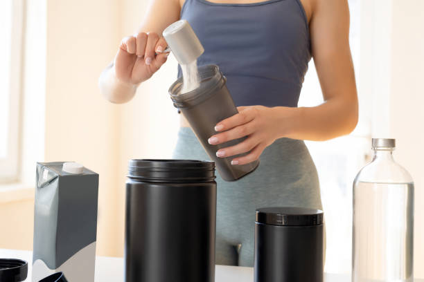 A woman preparing a protein shake in a shaker bottle, with protein powder containers and a water bottle on the counter.