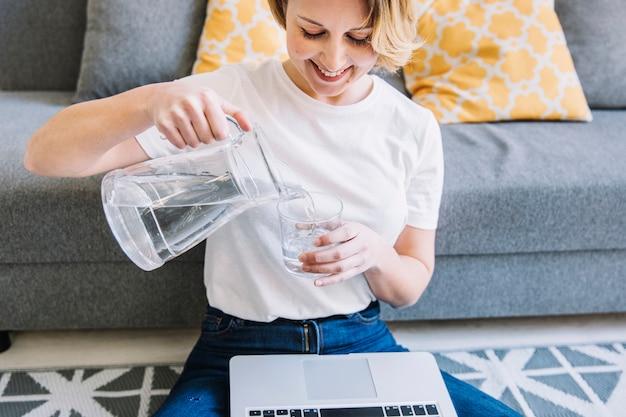 Smiling woman pouring water from pitcher into glass while sitting with laptop on sofa